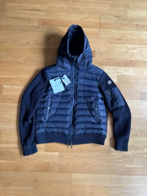 Blå Moncler cardigain  - Snygg mörkblå cardigain från Moncler med stickade ärmar och vadderad kropp. Jackan har huva, två dragkedjefickor fram och en ärmficka med Moncler-logga. Perfekt för kalla vinterdagar och riktigt trendig look.