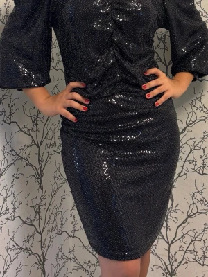 Svart glittrig kortklänning XS-M - Snygg svart kortklänning med glittrigt tyg och långa puffärmar. Klänningen har en figurnära passform och rynkade detaljer framtill som ger en cool look. Perfekt för dig som vill sticka ut med en trendig och festlig stil.