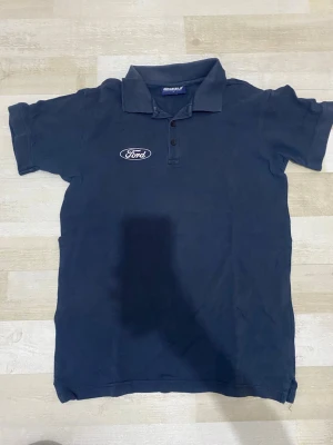 Mörkblå Ford pikétröja - Säljer en mörkblå pikétröja från Grizzly med Ford-logga broderad på bröstet. Klassisk krage och knäppning med tre knappar framtill. Perfekt för dig som gillar bilar och vill ha en enkel, stilren look.