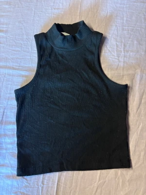 Svart ribbad ärmlös polotopp - Säljer en svart ribbad polotopp utan ärmar. Toppen har hög hals och är croppad, vilket ger en trendig och clean look. Perfekt att styla med jeans eller kjol för en cool vibe.