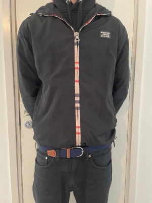 Burberry vindjacka  - Stilren svart vindjacka från Burberry med klassiskt rutigt mönster längs dragkedjan och liten logotyp på bröstet. Jackan har huva med dragsko, två sidofickor och justerbar nederkant. Perfekt för dig som vill ha en snygg och tidlös look.