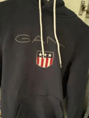 Mörkblå hoodie från GANT med logga - Mörkblå hoodie från GANT med vit dragsko i huvan och stor GANT-logga samt sköld med stjärnor och ränder på bröstet. Klassisk känguruficka framtill. Perfekt för en avslappnad och sportig stil.