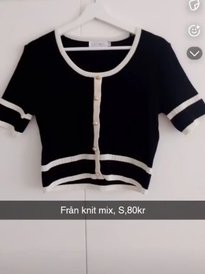 Svart kortärmad topp med pärlknappar - Kortärmad svart topp från Knit Mix med vita kontrastkanter och dekorativa pärlknappar framtill. Modellen är croppad och har rund halsringning samt ribbade detaljer vid ärmslut och nederkant. Perfekt för en trendig och chic look.
