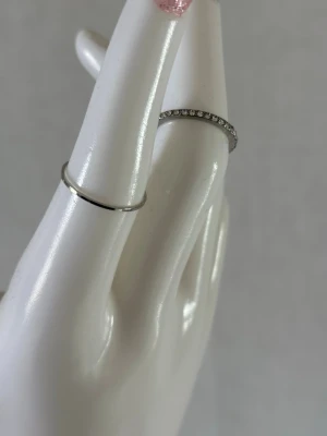 Två silverfärgade ringar - Säljer två stilrena ringar i silverfärg. Den ena ringen är enkel och slät medan den andra har små pärlliknande detaljer runt hela ringen. Perfekta att bära tillsammans eller var för sig för en minimalistisk look.