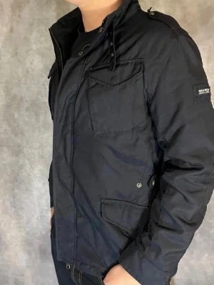 Woolrich jacka  - Snygg svart jacka från Woolrich med flera fickor framtill och hög krage. Jackan har dragkedja och knappar, samt en klassisk, rak passform. Perfekt för dig som vill ha en stilren och funktionell jacka till vardags. Jackan är i väldigt gott skick och inga defekter 