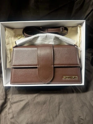 Jacquemus Bambino Bag  - Säljer nu min fina Jacquemus Bambino väska i brunt läder med guldfärgad text! Den är i helt ny skick, aldrig ens använd. Säljes pga felköp (skulle varit silver text istället) Storleken på väskan är 17,5 x 9 cm. (Medium size)  Låda, snören, dustbag medföljer✅  ✅Nyskick - helt oanvänd! (Frigolit kvar) ✅Låda, dustbag, snöre (allt og) medfölje ✅Självklart autentisk och äkta!  Vid fler frågor, tveka inte på att kontakta mig!