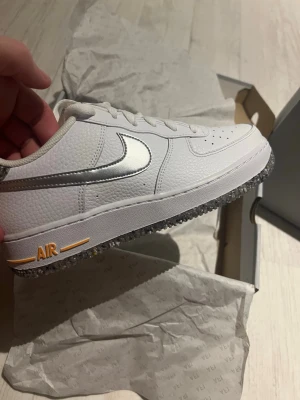 Nike Air Force 1 Low vit/silver - Säljer ett par Nike Air Force 1 Low i vitt läder med metallic silver swoosh och detaljer. Skon har klassisk perforerad tå, vit sula med orange AIR-text och genomskinlig yttersula med prickigt mönster. Perfekt för dig som vill ha en clean och stilren sneaker med unika detaljer. Har två par och har aldrig använt. 
