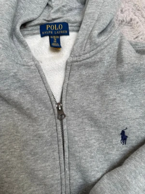 Grå hoodie från Polo Ralph Lauren - Klassisk grå hoodie med dragkedja från Polo Ralph Lauren. Tröjan har huva, fickor framtill och den ikoniska blå loggan broderad på bröstet. Tillverkad i mjuk bomullsblandning som känns skön mot huden. Perfekt för en avslappnad och stilren look.Knappt använd
