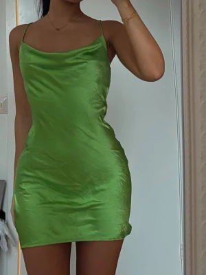 Grön satin kortklänning med tunna band - Snygg grön kortklänning i satin med tunna axelband och lätt draperad urringning. Klänningen har en figurnära passform och slät yta som ger en lyxig känsla. Perfekt för dig som vill sticka ut med en färgstark look.