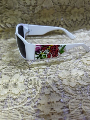 Vita solglasögon med blommigt mönster - Snygga vita solglasögon med rektangulära bågar och mörka glas. Sidorna har ett färgglatt blommigt mönster i rött, rosa och grönt som ger en unik och lekfull vibe. Perfekta för att sticka ut i solen! Helt ny 