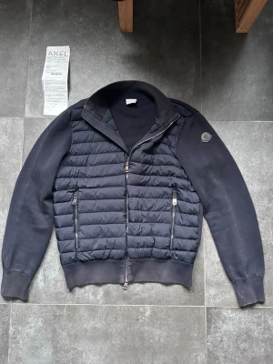 Moncler cardigan - Säljer en väldigt fin black label Moncler cardigan i rätt bra skick.  De enda anmärkningarna är på zip-upen och en liten anmärkning på framsidan av jackan på sista bilden. Men inget man tänker på när man har på sig den.  Marinblå i storlek L. Kvitto medföljer Bedöm skick själv utifrån bilderna.