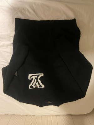 Svart stickad tröja med vit logga - Säljer en svart stickad tröja med en stor vit broderad logga på framsidan. Tröjan har långa ärmar och ribbade muddar vid ärmslut och nederkant. Perfekt för dig som gillar stilrena plagg med en twist.