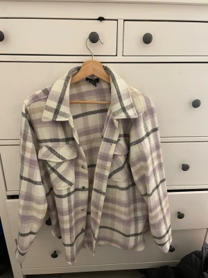 Rutig lila/vit kappa från Lindex - Snygg rutig kappa från Lindex i lila, vitt och svart. Oversized modell med stora bröstfickor och knappar framtill. Mjuk och skön, perfekt för lager på lager. Klassisk krage och långa ärmar. Passar dig som gillar trendiga och avslappnade looks.