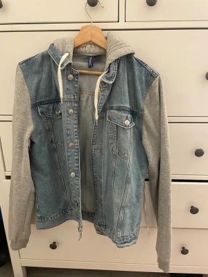Jeansjacka med grå luvtröjeärmar Divided - Snygg jeansjacka från Divided med ljusblå denim och grå sweatshirt-ärmar samt huva. Jackan har knappar framtill, två bröstfickor med lock och vita dragsnören i huvan. Perfekt för en avslappnad streetstil.