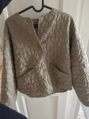 Beige quiltad cardigan från Zara - Säljer en beige quiltad cardiganjacka från Zara. Jackan har rund halsringning, tryckknappar framtill och två fickor. Materialet är vadderat med ett mönster av stora sömmar som ger en cool struktur. Perfekt för lager på lager och en avslappnad look.