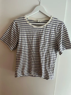 Randig t-shirt från Lindex - Säljer en klassisk randig t-shirt från Lindex i vitt och lila. T-shirten har korta ärmar, rund halsringning och är gjord i mjuk bomull. Perfekt till jeans eller shorts för en enkel och stilren look.