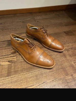 Bruna loafers i läder med broguemönster - Snygga bruna loafers i äkta läder med klassiskt broguemönster på tån och längs sömmarna. Skorna har låg klack, rundad tå och dekorativa perforeringar som ger en stilren och tidlös look. Perfekta för dig som vill ha en dressad men avslappnad stil. Oanvänd 