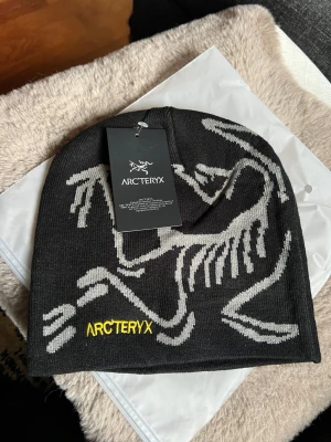 Svart mössa från Arc'teryx - Snygg svart mössa från Arc'teryx med grått skelettmönster och gul broderad logga framtill. Tillverkad i mjukt, stretchigt material som sitter skönt på huvudet. Perfekt för dig som vill ha en stilren och sportig look.