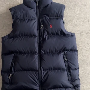 Svart dunväst från Polo Ralph Lauren - Snygg svart dunväst från Polo Ralph Lauren med röd broderad logga på bröstet. Västen har hög krage, dragkedja framtill och två fickor med dragkedja. Perfekt för lager på lager och riktigt stilren look.