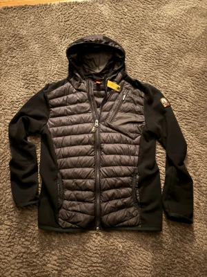 Parajumper hybrid  - Säljer en svart hybridjacka från Parajumpers i storlek L. Jackan har quiltad front och rygg, mjuka fleeceärmar och huva. Utrustad med dragkedja, bröstficka och snygg patch på ärmen. Perfekt för dig som vill ha både stil och funktion.