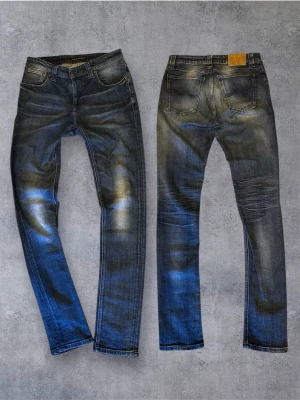 Selvage Nudie Jeans - Säljer ett par blå selvage jeans från Nudie Jeans med snygga slitningar och klassisk femficksmodell. Jeansen har en något tvättad look och är tillverkade i bomull. Perfekta för dig som gillar en modern och avslappnad stil.