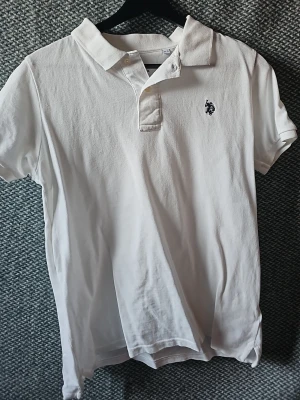 Vit pikétröja från U.S. Polo Assn - Klassisk vit pikétröja från U.S. Polo Assn med korta ärmar och broderad logga på bröstet. Tröjan har krage och knappar vid halsen. Tillverkad i mjuk bomull för en bekväm känsla och stilren look.