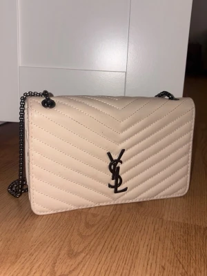 Beige quiltad YSL handväska - Säljer en beige quiltad handväska från Saint Laurent med klassisk YSL-logga i svart metall framtill. Väskan har kedjerem i mörk metall och elegant chevronmönster. Perfekt för dig som gillar stilrena och lyxiga accessoarer.