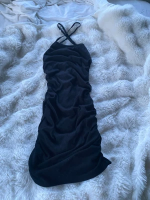 Svart klänning  - Snygg svart kort klänning med smala axelband som korsas i ryggen. Klänningen har en åtsittande passform och är lätt rynkad för en trendig look. Perfekt för dig som gillar stilrena och enkla plagg med en twist.