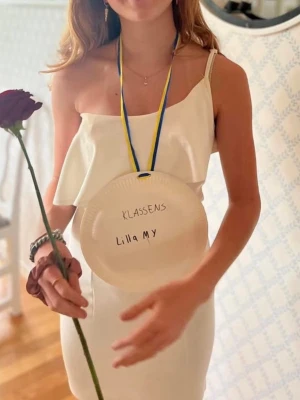 Vit klänning med volang - Snygg vit kortklänning med one shoulder och volangdetalj över bysten. Klänningen har en enkel och elegant design som passar perfekt till student, skolavslutning eller sommarens fester. 