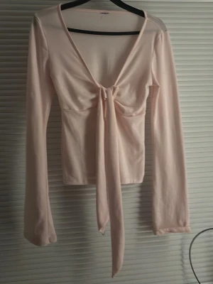 Ljusrosa långärmad topp med knyt - Trendig ljusrosa långärmad topp med djup v-ringning och knyt framtill. Toppen har en croppad passform och är tillverkad i mjukt bomullsmaterial. Perfekt för dig som gillar en feminin och stilren look.