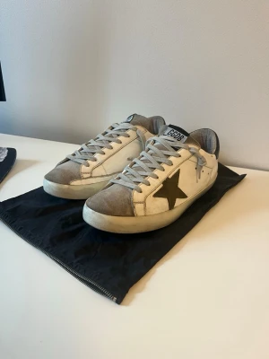 Golden Goose Superstar Skor - Säljer ett par vita Golden Goose sneakers med grå mockadetaljer och svart stjärna på sidan. Klassisk låg modell med GGDB-logga på plösen och slitna detaljer för en cool, avslappnad look. Skorna har gråa snören och gummisula. Väldigt fint skick samt så ingår dustabg, storlek 42.