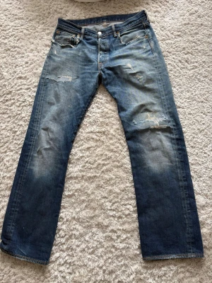 Blå slitna Levi's jeans straight fit - Säljer ett par blå Levi's jeans med straight fit och slitna detaljer på låren och knäna. Klassisk femficksmodell med knappgylf och läderpatch bak i midjan. Jeansen har en tvättad look och är tillverkade i bomull.W33 L32 men sitter mer som 31/32 i waist. Skriv vid funderingar eller frågor✌🏼