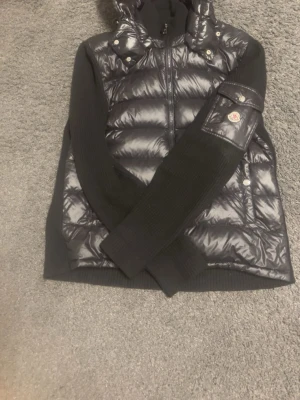 Svart moncler cardigan - Snygg svart dunväst från Moncler med glansig finish och stickade svarta ärmar. Västen har huva, dragkedja framtill och en ikonisk Moncler-logga på ärmen. Perfekt för lager-på-lager och streetwear-vibe. Materialet är en mix av dun och stickat tyg.