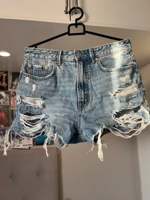 Blå slitna jeansshorts Vintage - Säljer ett par blå denimshorts från Vintage med hög midja och rejält slitna detaljer framtill och baktill. Shortsen har fransiga benslut, klassiska fem fickor och knappgylf. Perfekta för en avslappnad och trendig sommarlook.