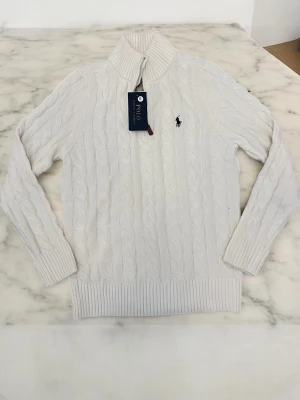 Vit quarter zip tröja Polo Ralph Lauren - Kabelstickad vit tröja från Polo Ralph Lauren med hög krage och dragkedja. Tröjan har ribbade muddar och nederkant samt den klassiska loggan broderad på bröstet. Perfekt för dig som gillar stilrena och tidlösa plagg.