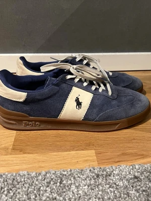 Blåa Polo Ralph Lauren sneakers - Snygga sneakers från Polo Ralph Lauren i blå mocka med vita detaljer och bruna gummisulor. Klassisk låg modell med vita snören och Polo-logga på sidan samt bak på hälen. Skitsnyggt att har nu till sommaren! Kan starkt rekommendera dessa. Original pris ligger på 1600kr