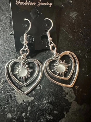 Silvriga hjärtörhängen med solmotiv - Ett par silvriga örhängen med öppna hjärtan och ett solformat hänge i mitten. Örhängena har krokar och är tillverkade i metall. Perfekta för dig som gillar unika och trendiga accessoarer med cool design.