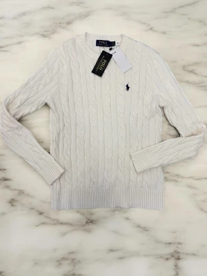 Vit kabelstickad tröja Polo Ralph Lauren - Kabelstickad vit tröja från Polo Ralph Lauren med rund halsringning och lång ärm. Tröjan har den klassiska broderade loggan på bröstet och ribbade muddar vid ärmslut och nederkant. Perfekt för en stilren och tidlös look.