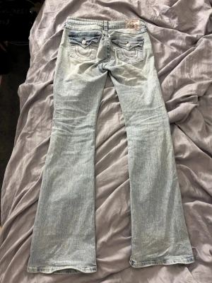 Ljusblå bootcut jeans True Religion - Säljer ett par ljusblå bootcut jeans från True Religion med ikoniska bakfickor och röda kontrastsömmar. Jeansen har ficklock med knapp och klassisk etikett bak i midjan. Perfekta för dig som gillar en avslappnad men trendig stil.