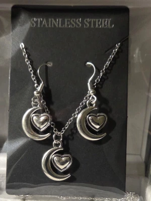 Smyckesset med måne och hjärta - Snyggt smyckesset i rostfritt stål med halsband och örhängen. Både halsbandet och örhängena har silverfärgade hängen i form av en måne med ett hjärta i mitten. Perfekt för dig som gillar unika detaljer och stilrena accessoarer.