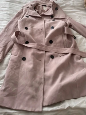 Ljusrosa trenchcoat med bälte - Snygg ljusrosa trenchcoat med klassisk dubbelknäppning och matchande bälte i midjan. Jackan har svarta knappar, bred krage och två fickor framtill. Perfekt för dig som vill ha en stilren och trendig look.