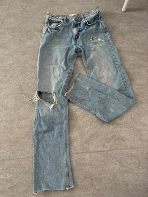 ZARA Ripped Jeans, storlek 36 - Säljer ett par riktigt snygga och trendiga ljusblå jeans från Zara i storlek EUR 36 (USA 4 / MEX 26).  De har en cool look med ett större hål på ena knät och slitna benslut. Jeansmodellen är raka i benen och är i fint skick! ⚡️⚡️  Våga pruta <3