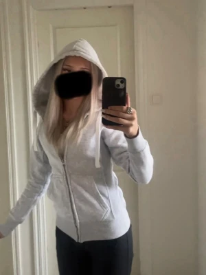 Ljusgrå zip hoodie med huva russemerch  - Snygg ljusgrå hoodie med dragkedja framtill och stor huva. Tröjan har vita dragsnören, ribbade muddar och två fickor på magen. Den har tappat lite färg på dragkedjan.