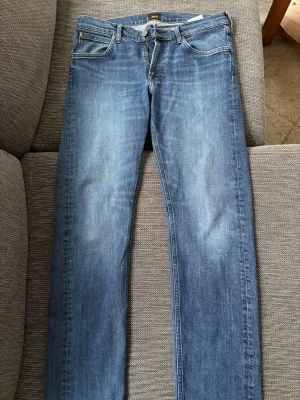 Blå raka Lee jeans  - Säljer ett par blå jeans med rak passform och klassisk tvätt. Jeansen har fem fickor, normal midja och subtila slitningar framtill för en avslappnad look. Perfekta till vardagsstilen och tillverkade i mjukt denimtyg.