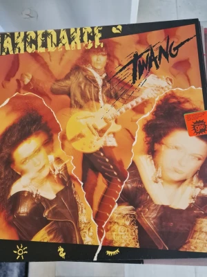 Twang vinylskiva från TranceDance - Vinylskiva med albumet 'Twang' av TranceDance. Omslaget har en orange och svart design med bilder på bandmedlemmar och elgitarr. Baksidan listar låtar som 'Money can't buy' och 'Heaven in your eyes'. Skivan är utgiven av CBS Records och har en tydlig retrostil. Inga repor. Välvårdad.