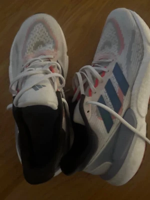 Vita Adidas sneakers med blå ränder - Snygga vita Adidas sneakers med klassiska blå och rosa ränder på sidan. Ovandelen är i mesh för bra andningsförmåga och sulan är tjock och dämpad för extra komfort. Skorna har vita snören och svarta detaljer vid hälen och plösen.