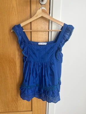Supersöt blå broderad topp från MNG - Supersöt blå topp från MNG med broderade detaljer och volangkant. Toppen har breda axelband, smock upptill och fina spetsdetaljer längs nederkanten. Perfekt för varma dagar och ger en fräsch look till din outfit.
