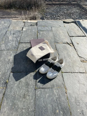 Valentino vita sneakers med olivegrönt band - Snygga vita sneakers från Valentino med ett brett olivgrönt band över vristen och diskret logga på plösen. Skorna har klassisk låg profil och är tillverkade i läder. Kommer med originalkartong och dustbag.