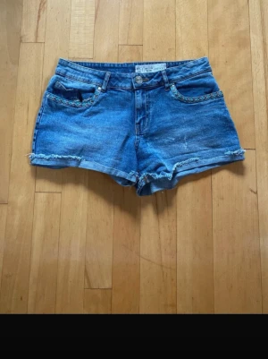 Blå jeansshorts med fransig kant - Säljer ett par blå jeansshorts med fransig kant och flätad detalj vid fickorna. Klassisk femficksmodell med knapp och dragkedja framtill. Perfekta för varma dagar och enkel att matcha med allt.