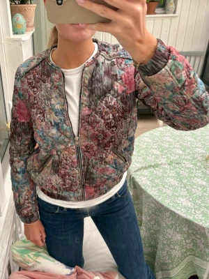 Mönstrad quiltad bomberjacka - Säljer en unik quiltad bomberjacka med blommigt mönster i vinröda, blå och gröna toner. Jackan har dragkedja framtill, ribbade muddar och två fickor. Perfekt trendig inför våren!🌼🌸